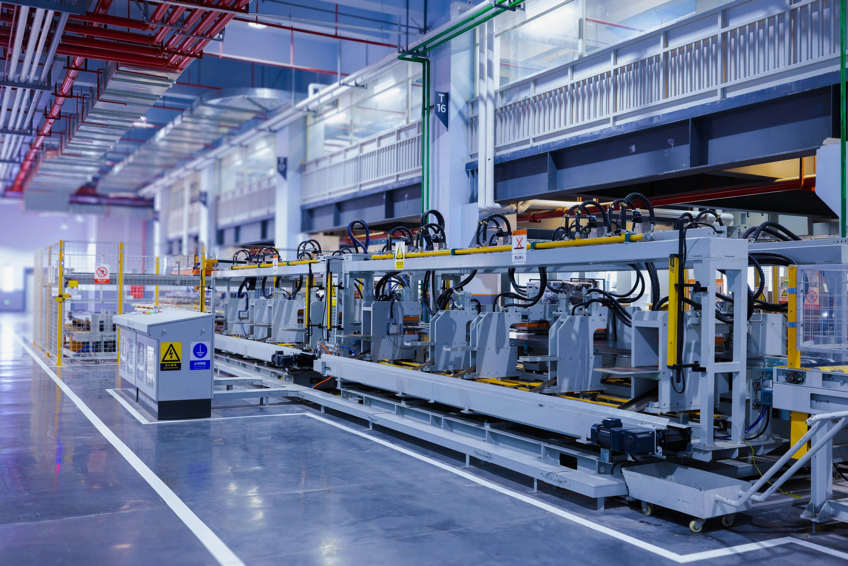 Automatisierte Umformlinie in einer hellen Werkhalle: Stationen mit Hydraulikschläuchen in einer Reihe, davor ein Schaltschrank und gelbe Schutzgitter