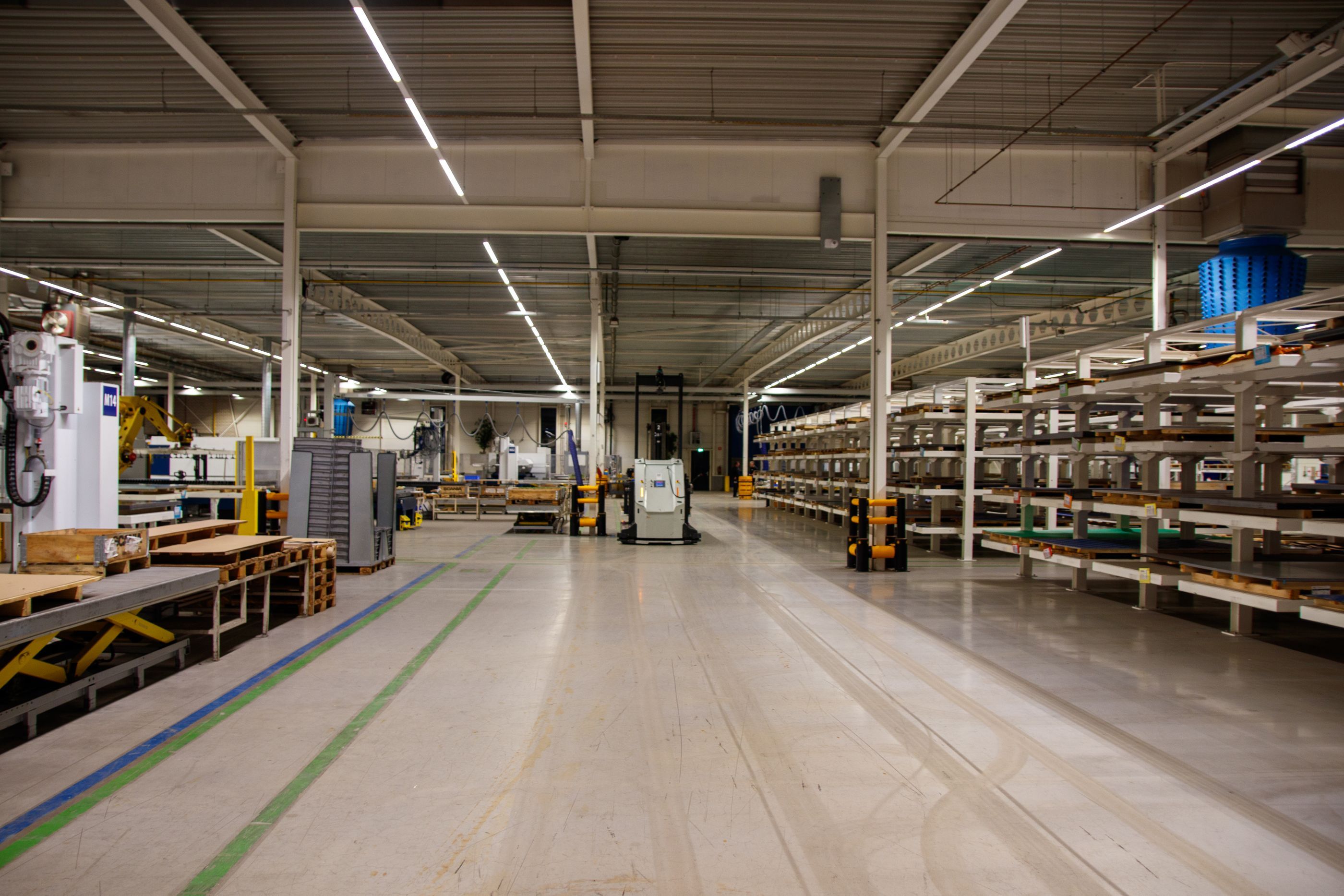 Blick durch eine Produktionshalle: links Maschinen und Paletten, rechts Regale, darüber LED-Lichtbänder unter der Hallendecke